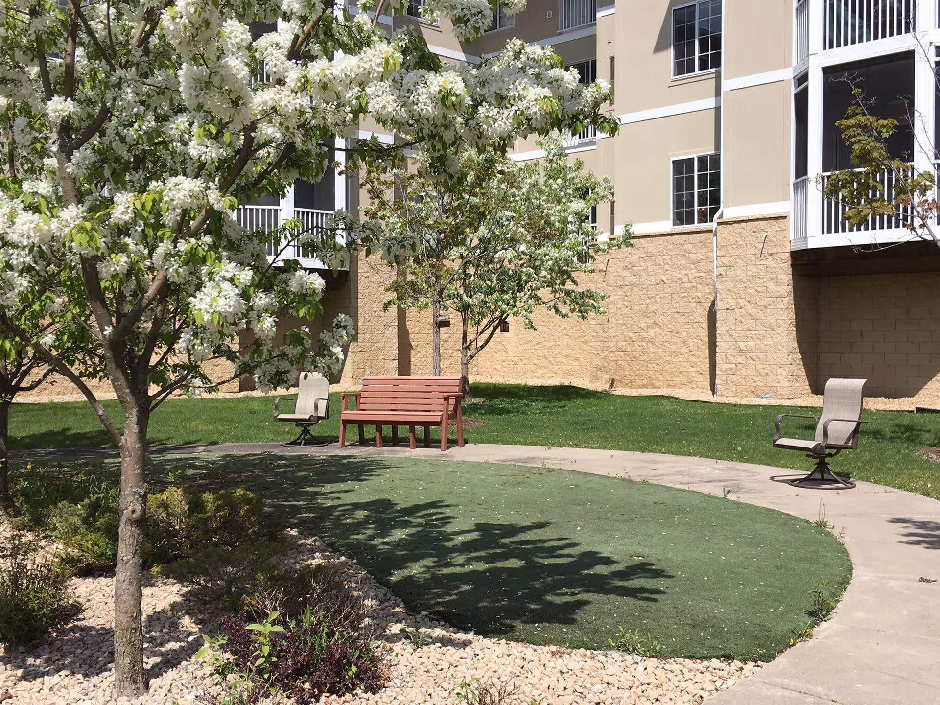 Outdoor garden area with blooming white-flowered trees, a curved concrete pathway, a red wooden bench, and two beige swivel chairs in front of a beige building with balconies.