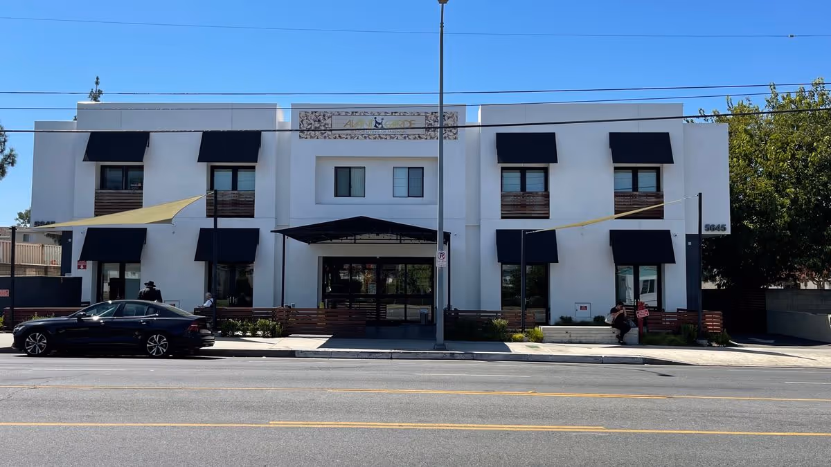 Front exterior of a two-story white senior living building with black awnings, an entrance canopy, parked car and people on the sidewalk.
