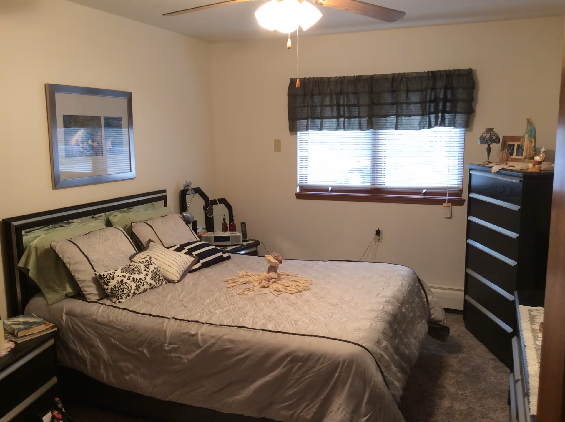 A bedroom with a neatly made bed featuring multiple pillows and a decorative towel arrangement on top. The room has a window with blinds and a dark valance, a ceiling fan with a light, a dresser with various items on top, and a nightstand with a lamp and other small objects. The walls are plain and light-colored.