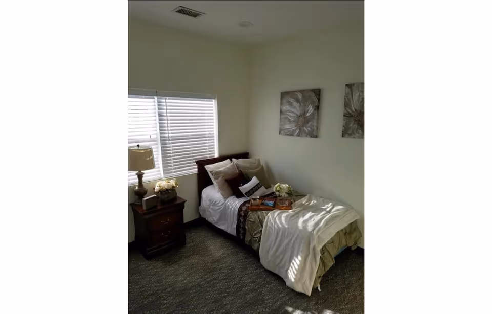 Sunlit single bedroom with a made twin bed, nightstand with a lamp and flowers, and two floral wall art pieces.