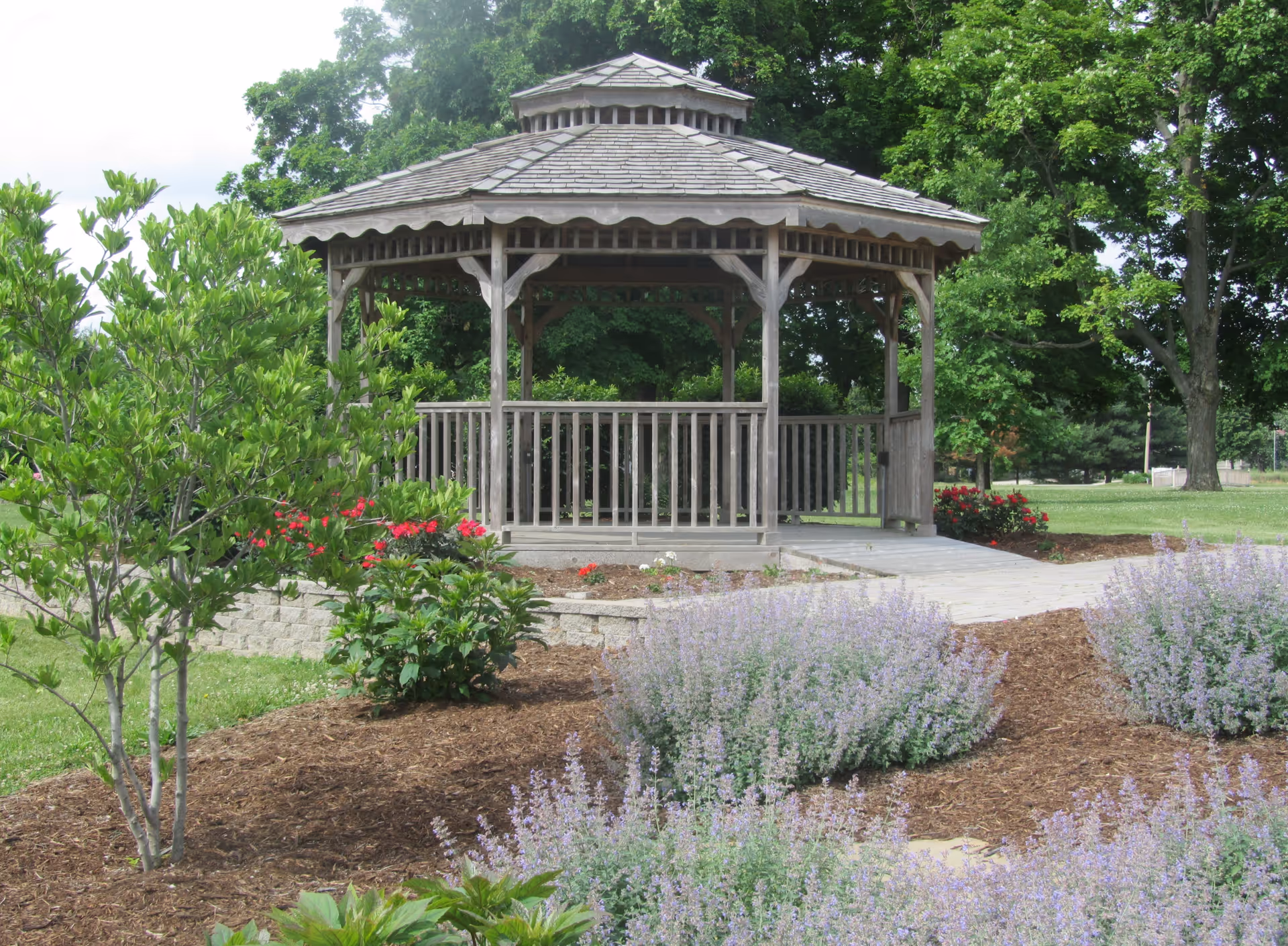 A wooden gazebo with a shingled roof situated in a landscaped garden area with various green shrubs, flowering plants, and trees in the background under a partly cloudy sky.