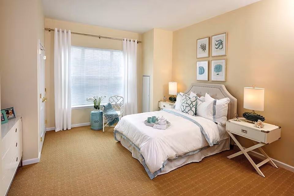 A bright and cozy bedroom with beige walls and carpet. The room features a neatly made bed with white and light blue bedding, two bedside tables each with a lamp, and four framed artworks above the bed. A window with white curtains allows natural light to fill the room. There is a small chair with a throw blanket and a decorative blue ceramic stool near the window.