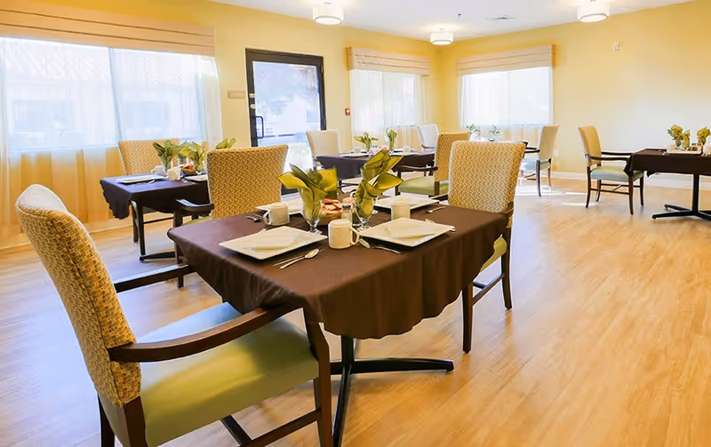 Bright dining room with multiple tables set for meals, upholstered chairs, and potted centerpieces.