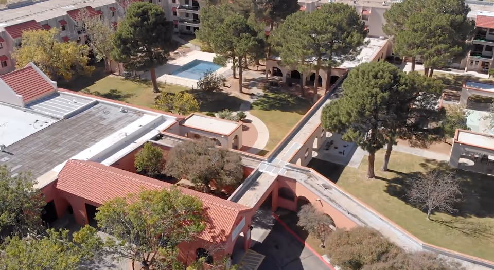 Aerial view of Good Samaritan Society White Acres showing a courtyard with trees, grass, walkways, and a swimming pool surrounded by buildings with red-tiled roofs.