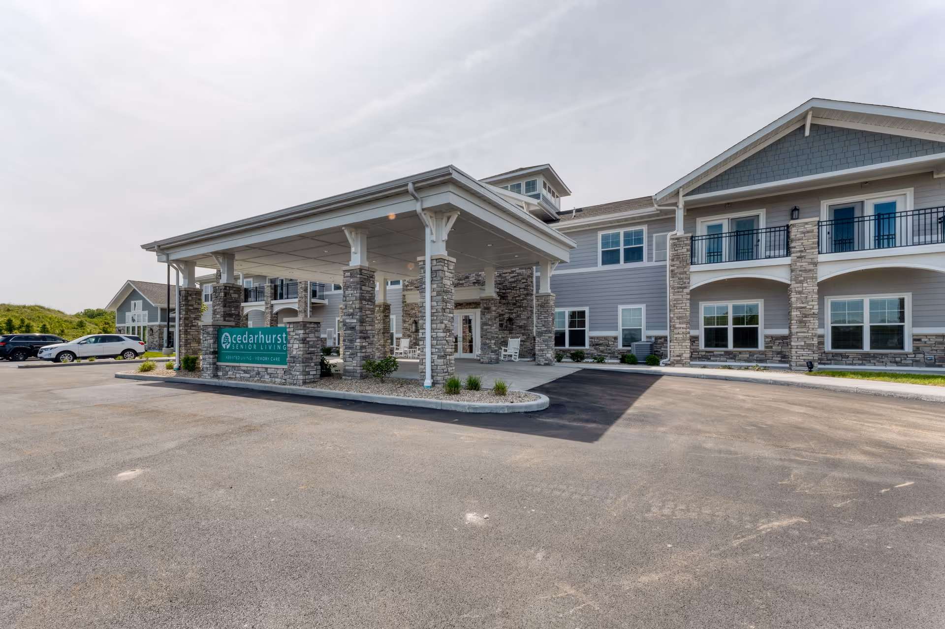 Exterior view of Cedarhurst Senior Living of McHenry building with a covered entrance supported by stone pillars, a parking lot in front, and a clear sky above.