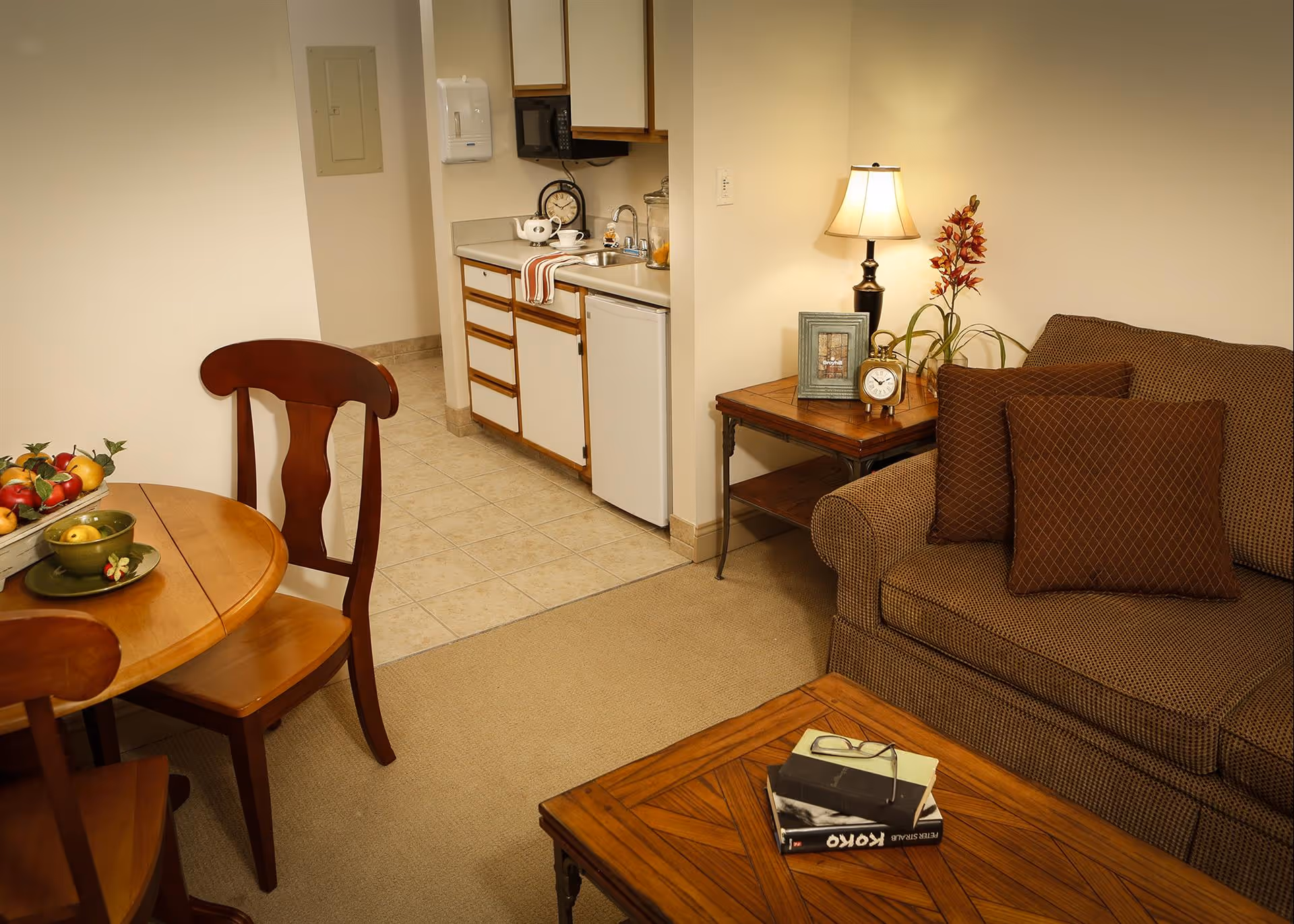 A cozy living area with a brown patterned sofa adorned with matching pillows, a wooden coffee table with books and glasses on it, a side table with a lamp, a clock, a framed picture, and a flower vase. Adjacent to the living area is a small kitchenette with white cabinets, a mini fridge, a microwave, and a sink. A wooden dining table with chairs and a bowl of fruit is partially visible.