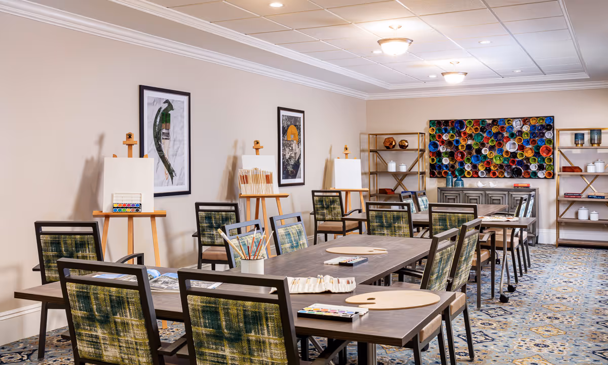 A well-lit art activity room with several tables and chairs arranged for group painting sessions. The tables have paint palettes, brushes, and blank canvases on easels. The walls are decorated with framed artwork and a colorful circular wall art piece. Shelves with decorative items and books are also visible.