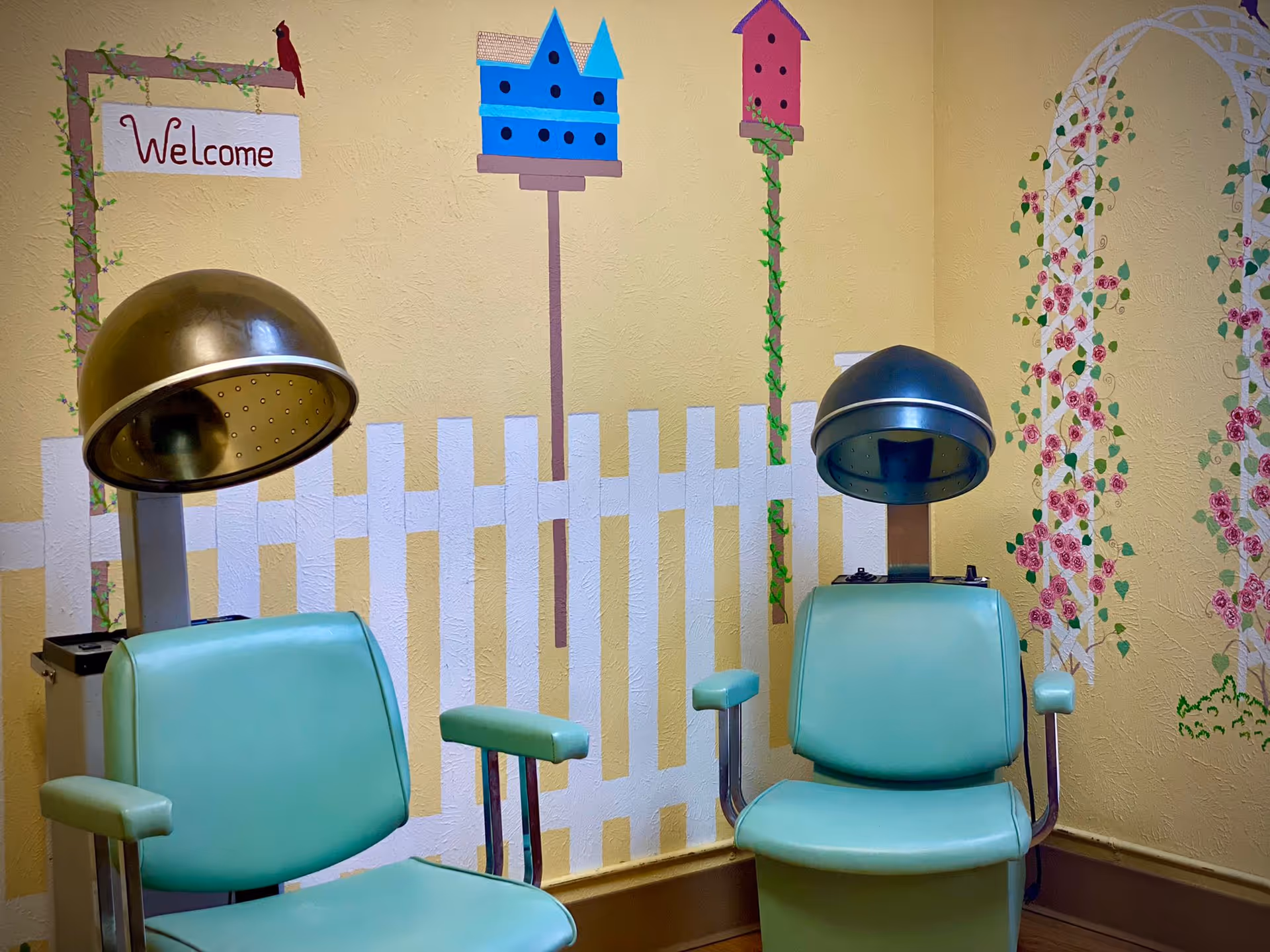 Two vintage green salon chairs with attached hair dryers in a room with yellow walls decorated with a white picket fence mural, birdhouses, flowers, and a welcome sign.