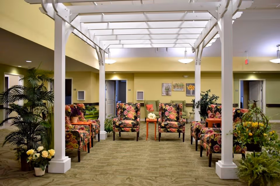A brightly lit indoor seating area with floral patterned armchairs arranged in a semi-circle under a white pergola structure. The room has light yellow walls, green carpeting, potted plants, and framed floral artwork on the back wall.