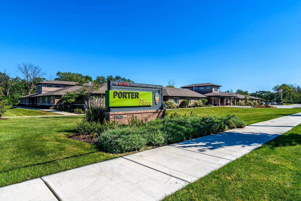 Exterior view of Porter Place, a single-story building with a well-maintained lawn, shrubs, and a sidewalk leading to the entrance under a clear blue sky. A large sign in front reads 'Porter Place' with additional text indicating it is an Anthem Memory Care Community.