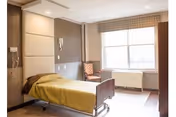 A simple, clean hospital-style room with a single bed covered with a yellow blanket, a chair near the window, and a large window letting in natural light. The walls are neutral colored, and there is a call button and telephone mounted on the wall near the bed.