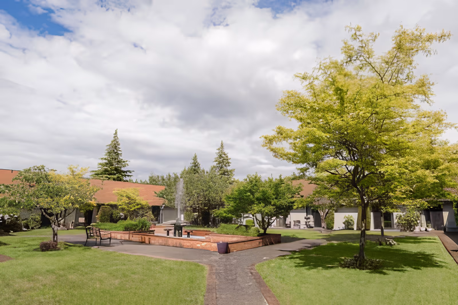 Courtyard with a central fountain, benches, trees, and surrounding single-story buildings under a cloudy sky.