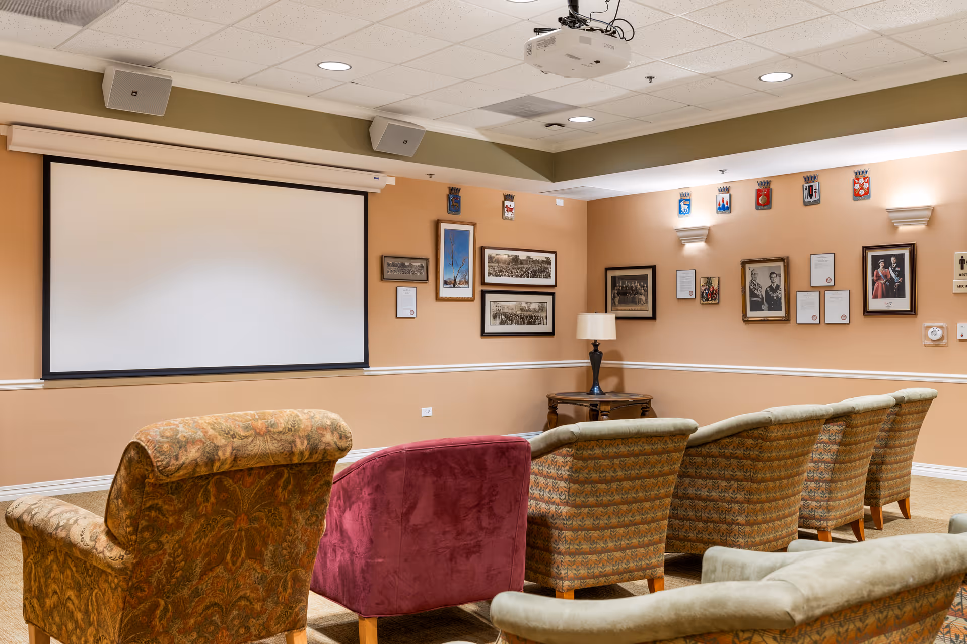 A cozy room with a projector screen on the wall, several upholstered armchairs arranged in rows facing the screen, framed pictures and plaques on the walls, and a small table with a lamp in the corner.