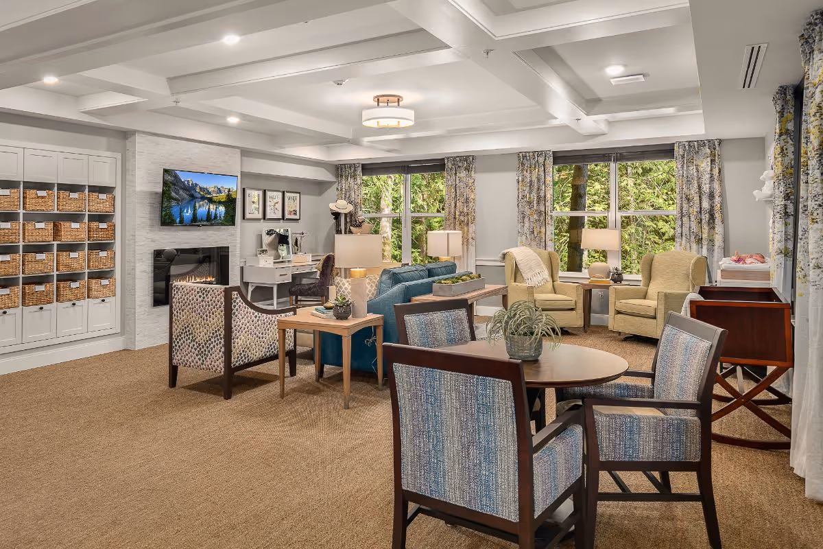 Bright furnished communal living room with chairs, tables, a fireplace and TV, and large windows showing trees.