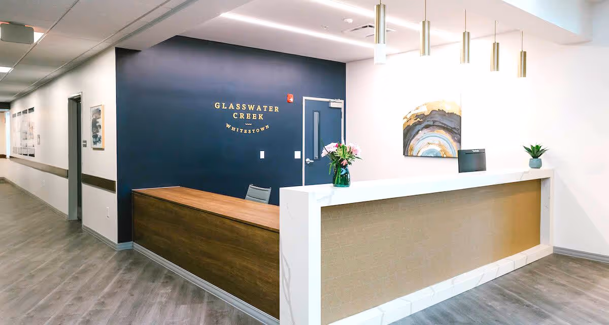 Reception area of Glasswater Creek of Whitestown featuring a modern front desk with a wooden and white marble finish, a vase with flowers, a small potted plant, pendant lights hanging from the ceiling, and a dark blue accent wall with the facility name in gold letters.