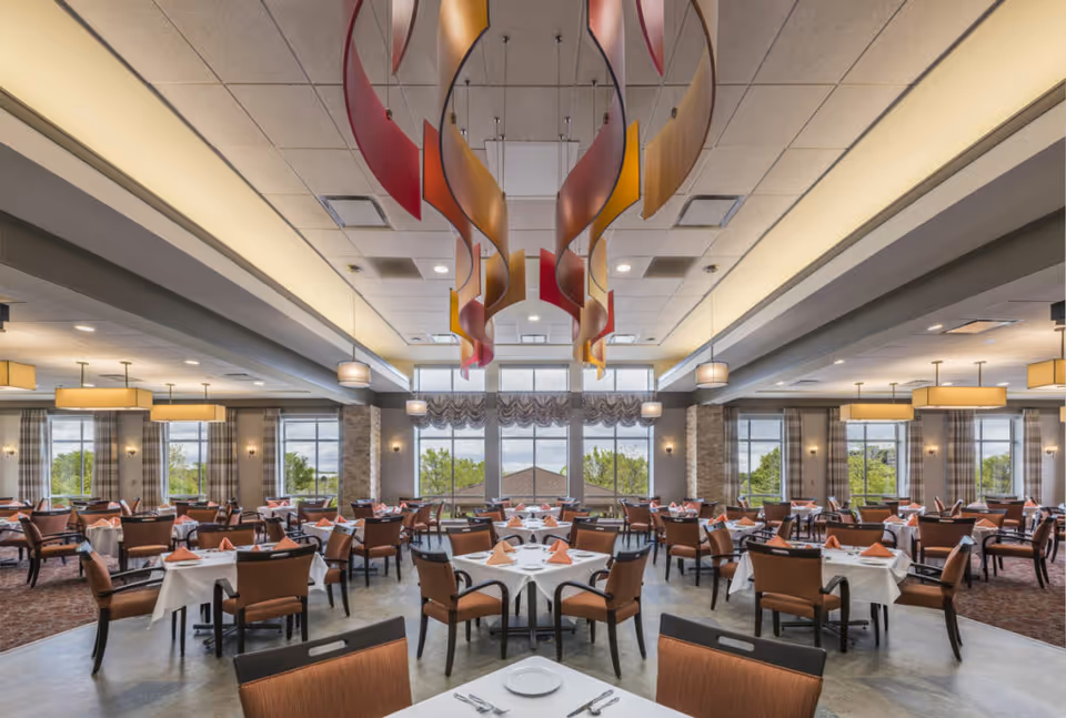 Spacious, well-lit dining room with multiple tables set with white linens and folded napkins, decorative ceiling sculptures, and large windows.