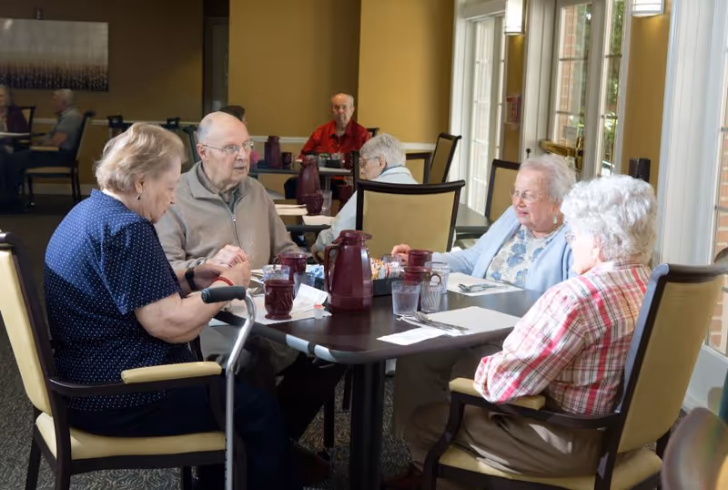 A group of elderly people sitting around a dining table in a well-lit room with large windows. The table has pitchers, glasses, and utensils, and the individuals appear to be engaged in conversation or waiting for a meal. Additional elderly people are visible in the background seated at other tables.
