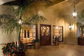 Exterior entrance of a building at night with a wooden door featuring diamond-shaped window panes, flanked by two large windows. The area is decorated with potted plants and illuminated by wall-mounted lantern-style lights and hanging pendant lights.