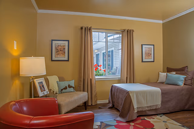 Cozy bedroom with a single bed, seating (red chair and bench), a lamp, and a window with curtains.