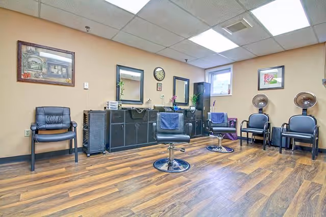 Interior view of a salon area in a senior living facility with two black salon chairs in front of mirrors and cabinets. There are additional black chairs along the walls, framed pictures, a clock, and wood-patterned flooring.