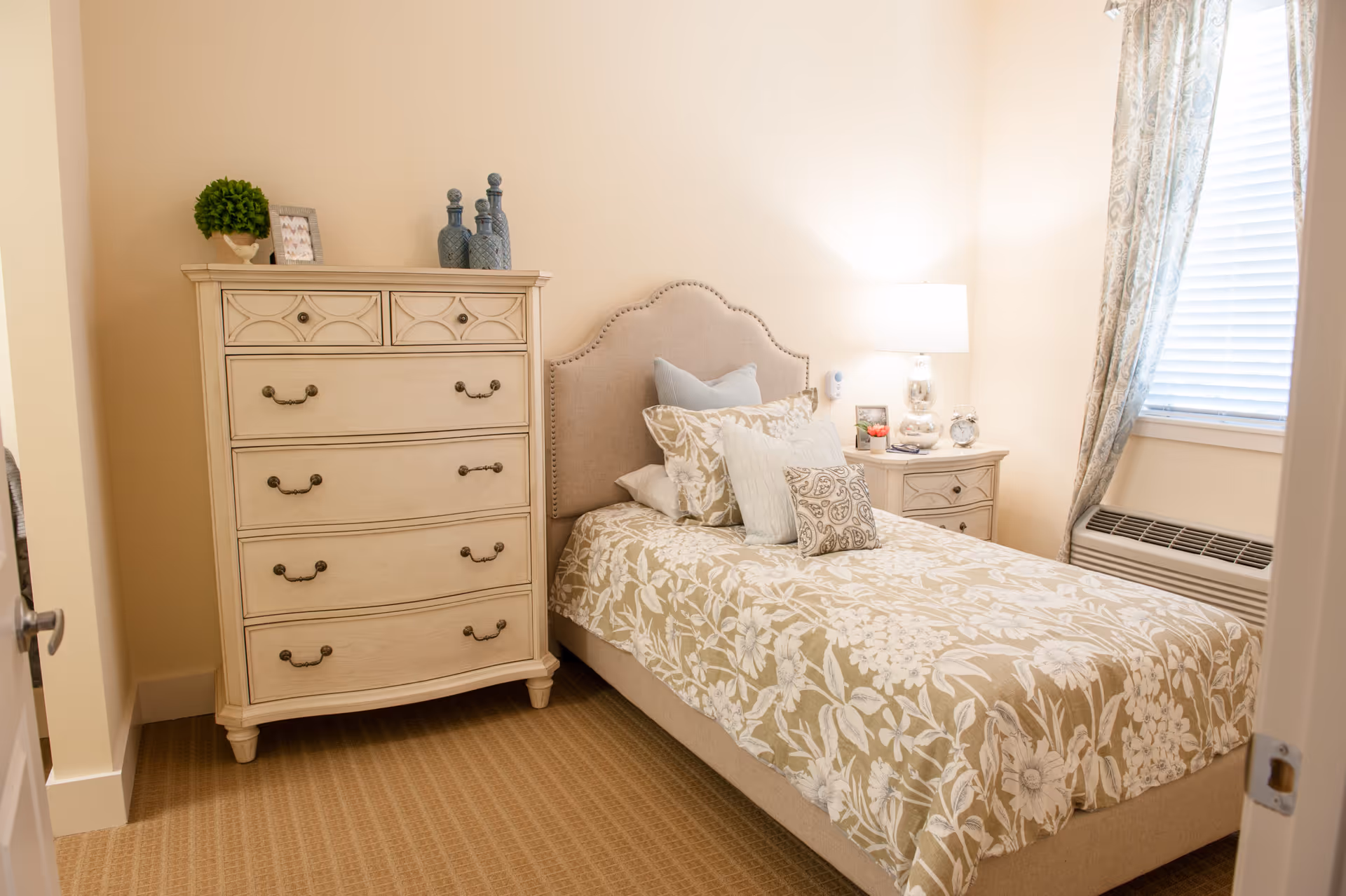 A cozy bedroom with a single bed featuring a beige floral bedspread and multiple pillows. Next to the bed is a nightstand with a lamp, a small plant, a clock, and a framed photo. On the left side of the room is a tall dresser with decorative items on top, including a small green plant and blue glass bottles. A window with patterned curtains and blinds is on the right wall, and there is a heating/cooling unit below the window.