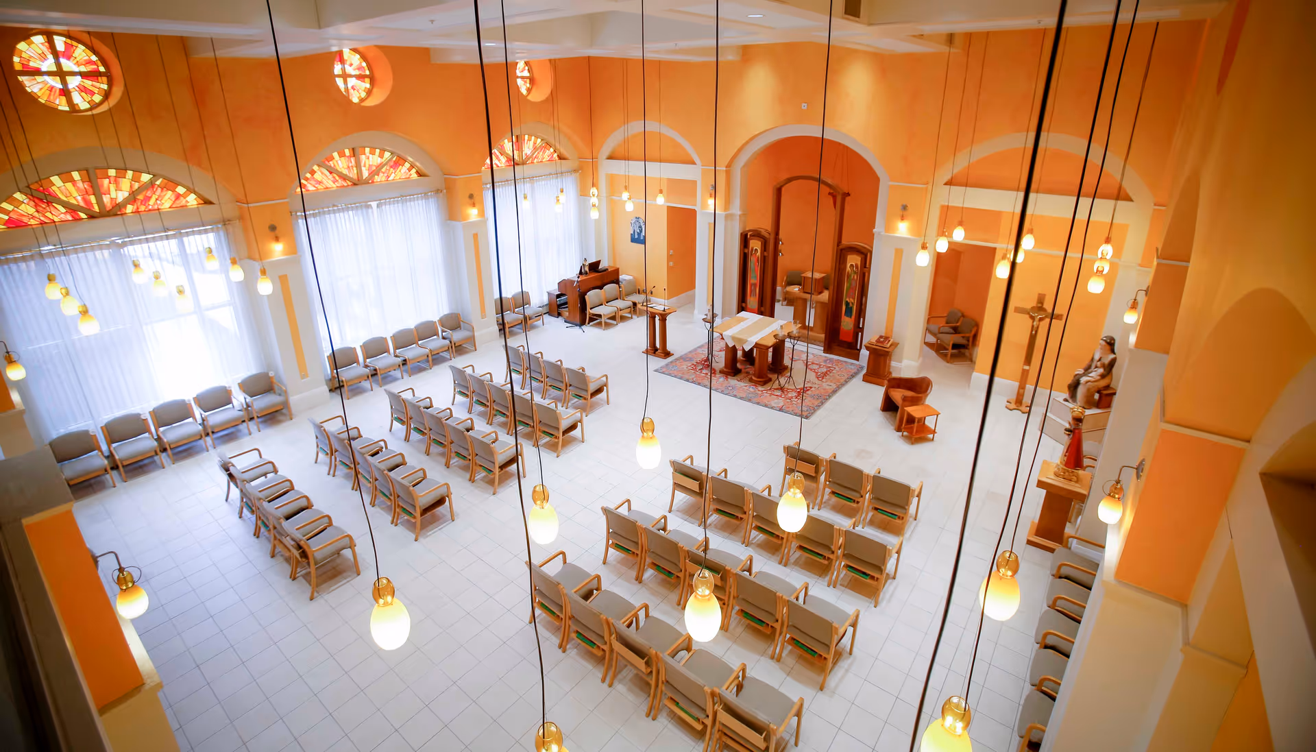 Interior view of a chapel or worship room with rows of wooden chairs arranged facing an altar area. The room has high ceilings with hanging pendant lights and large windows with stained glass designs near the ceiling. The walls are painted in warm orange tones and there are religious statues and a cross near the altar.