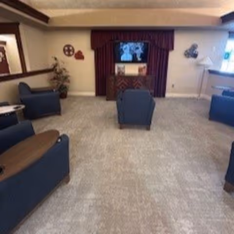 A cozy living room area with blue upholstered chairs arranged around a carpeted floor. A television is mounted on the wall above a wooden cabinet with a burgundy curtain backdrop. The room has neutral-colored walls with some decorative wall hangings and a small lamp on a side table near a window.