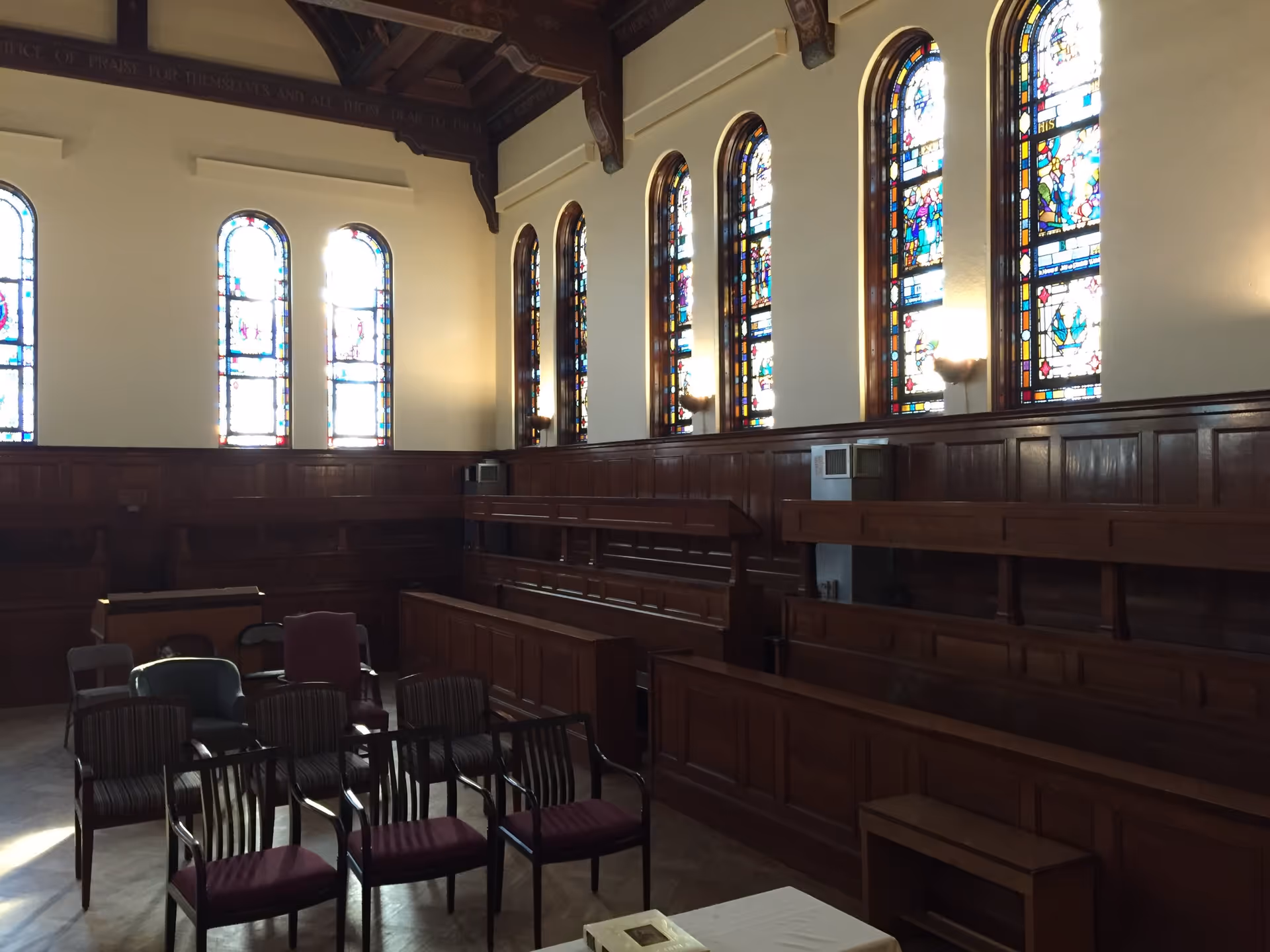 Interior of a chapel or worship room with wooden pews and chairs arranged facing a table. The room features tall stained glass windows along the wall and wooden paneling on the lower half of the walls. The ceiling has decorative wooden beams.
