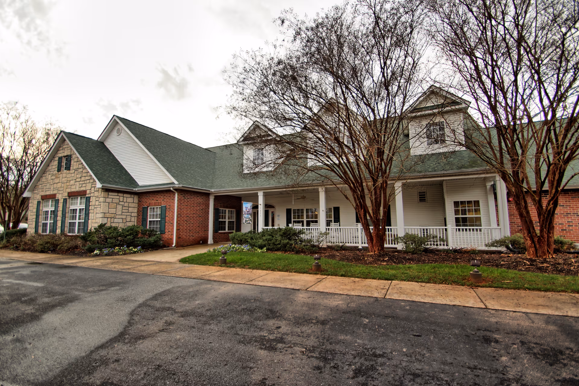 Exterior view of a single-story building with a green roof, brick and stone facade, white porch with rocking chairs, and leafless trees in front. The sky is cloudy and the pavement is wet.