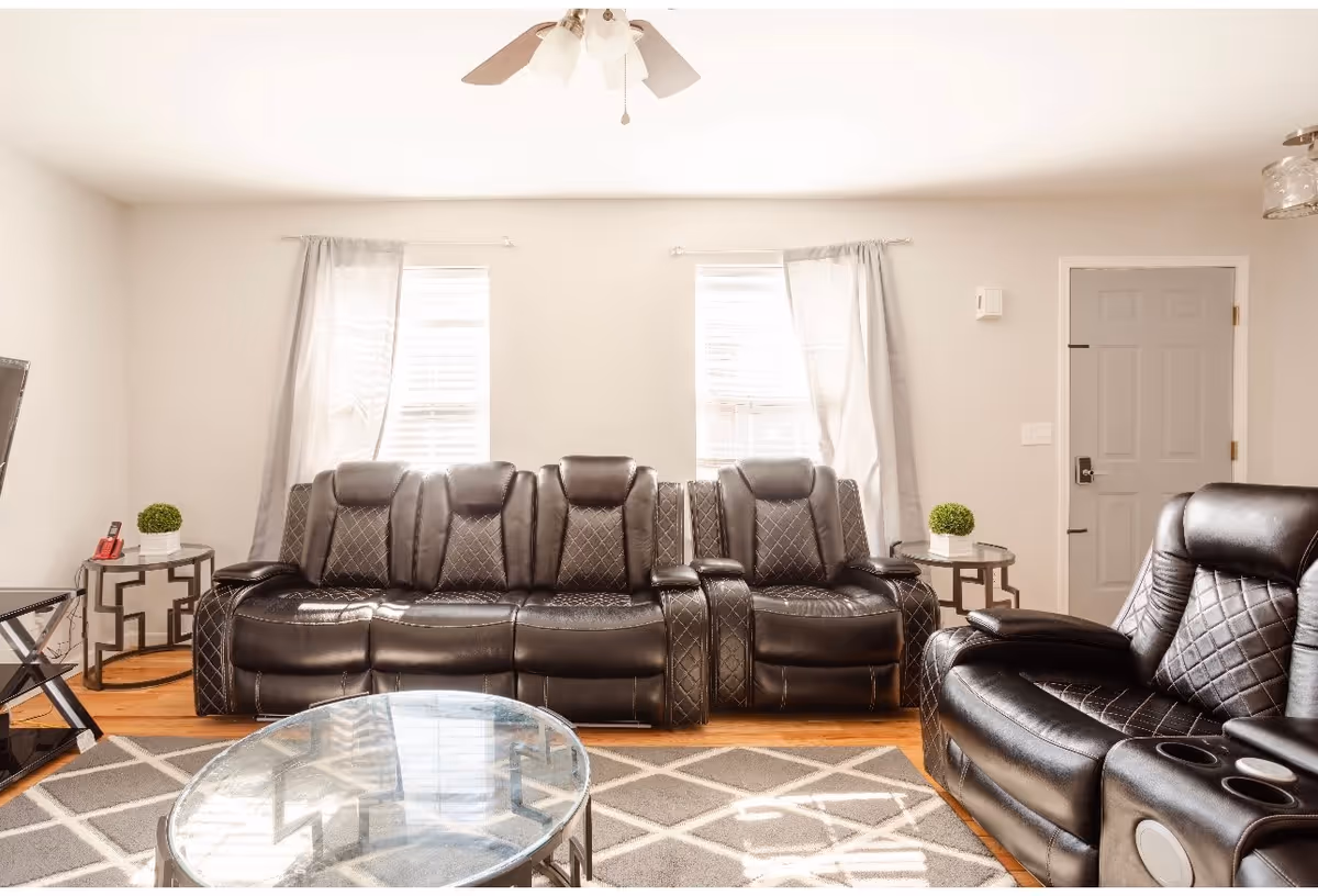 A bright living room with two large black leather recliner sofas, a glass coffee table on a patterned rug, two small side tables each with a small green plant, two windows with light curtains, a ceiling fan, and a closed gray door.