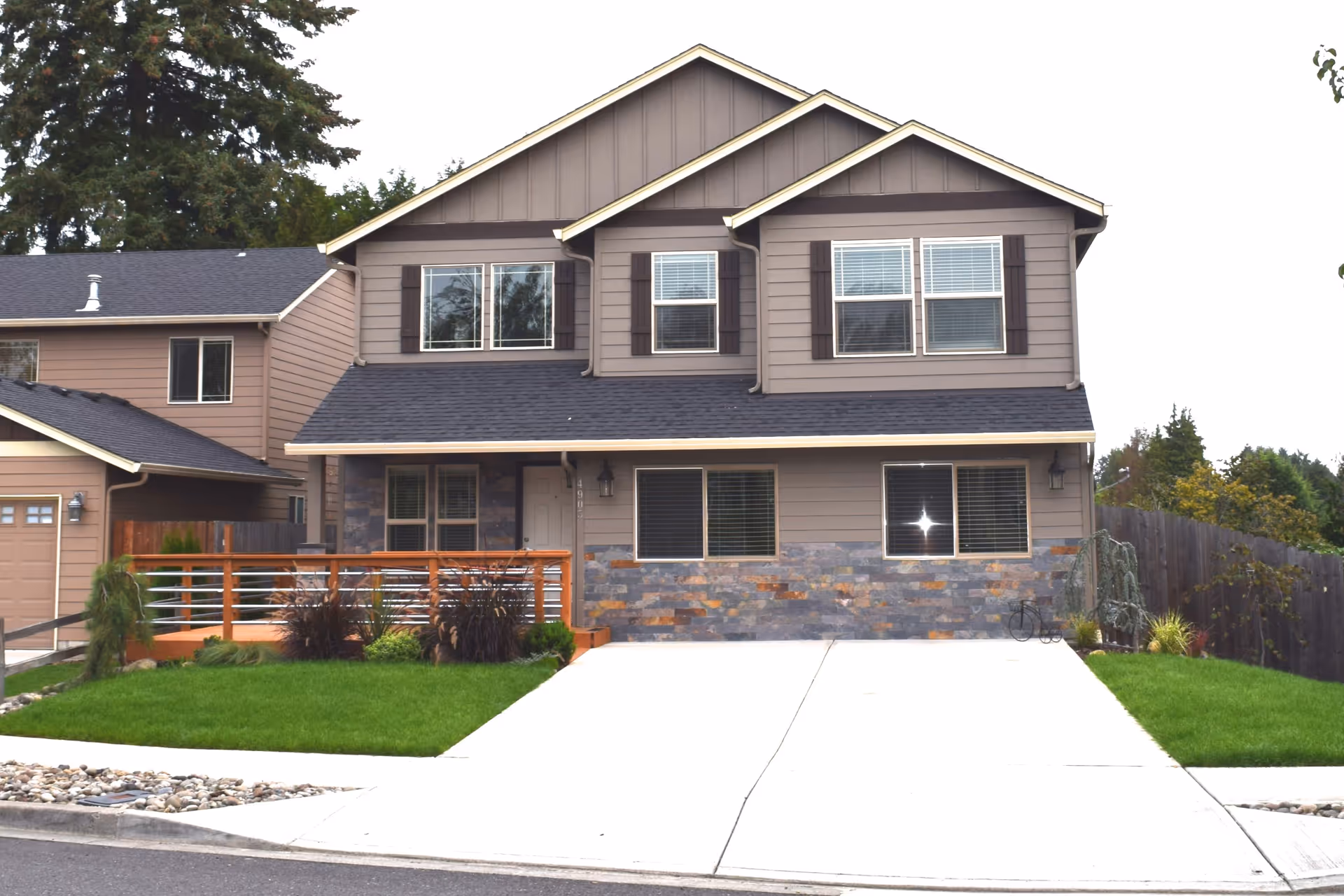 Front exterior view of a two-story residential building with gray siding, dark shutters, and a stone accent on the lower part of the facade. The house has a black shingled roof, several windows with white blinds, a small porch with wooden railing, and a concrete driveway leading up to the entrance. There is a well-maintained green lawn with some shrubs and plants on either side of the driveway.