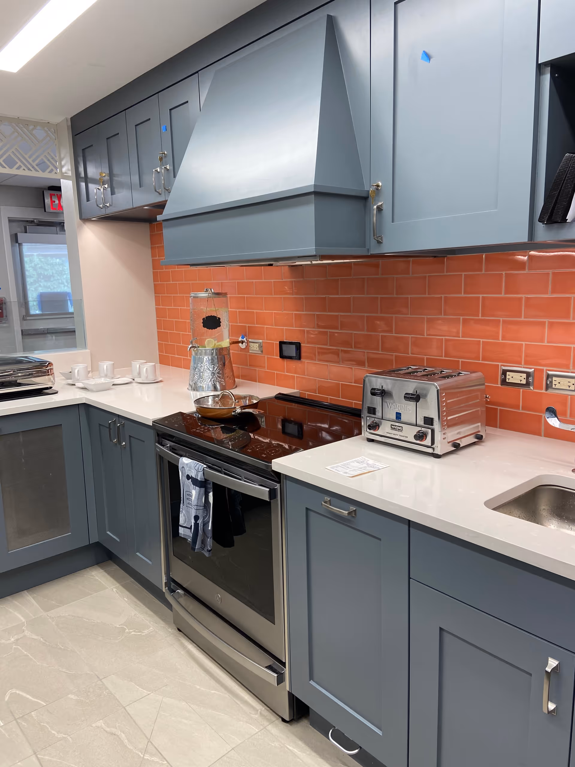 Modern kitchen with blue-gray cabinets, a stainless steel stove with a towel hanging on the handle, a silver toaster, a glass beverage dispenser with lemon water, and a sink. The backsplash is made of orange subway tiles, and the floor is light-colored tile.