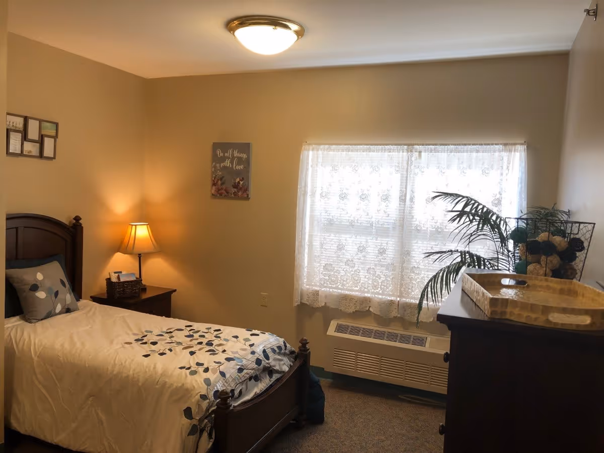 A cozy bedroom with a single wooden bed featuring a white and blue patterned comforter and a decorative pillow. There is a wooden nightstand with a lit lamp and a basket on it. A window with lace curtains allows natural light into the room. A wooden dresser with a decorative tray and a basket of decorative balls sits against the opposite wall. A framed wall art with the phrase 'Do all things with love' is hung above the nightstand.