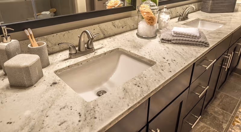 Close-up view of a bathroom countertop with two sinks, silver faucets, a large mirror, and various bathroom accessories including toothbrush holders, jars with cotton balls and sponges, and folded towels.