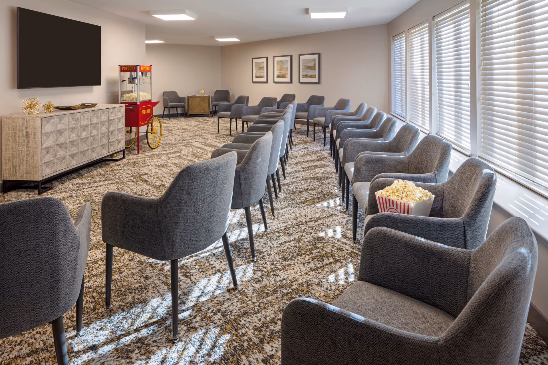 A bright communal media room with rows of gray chairs facing a wall-mounted TV, a popcorn cart, and large windows with blinds.