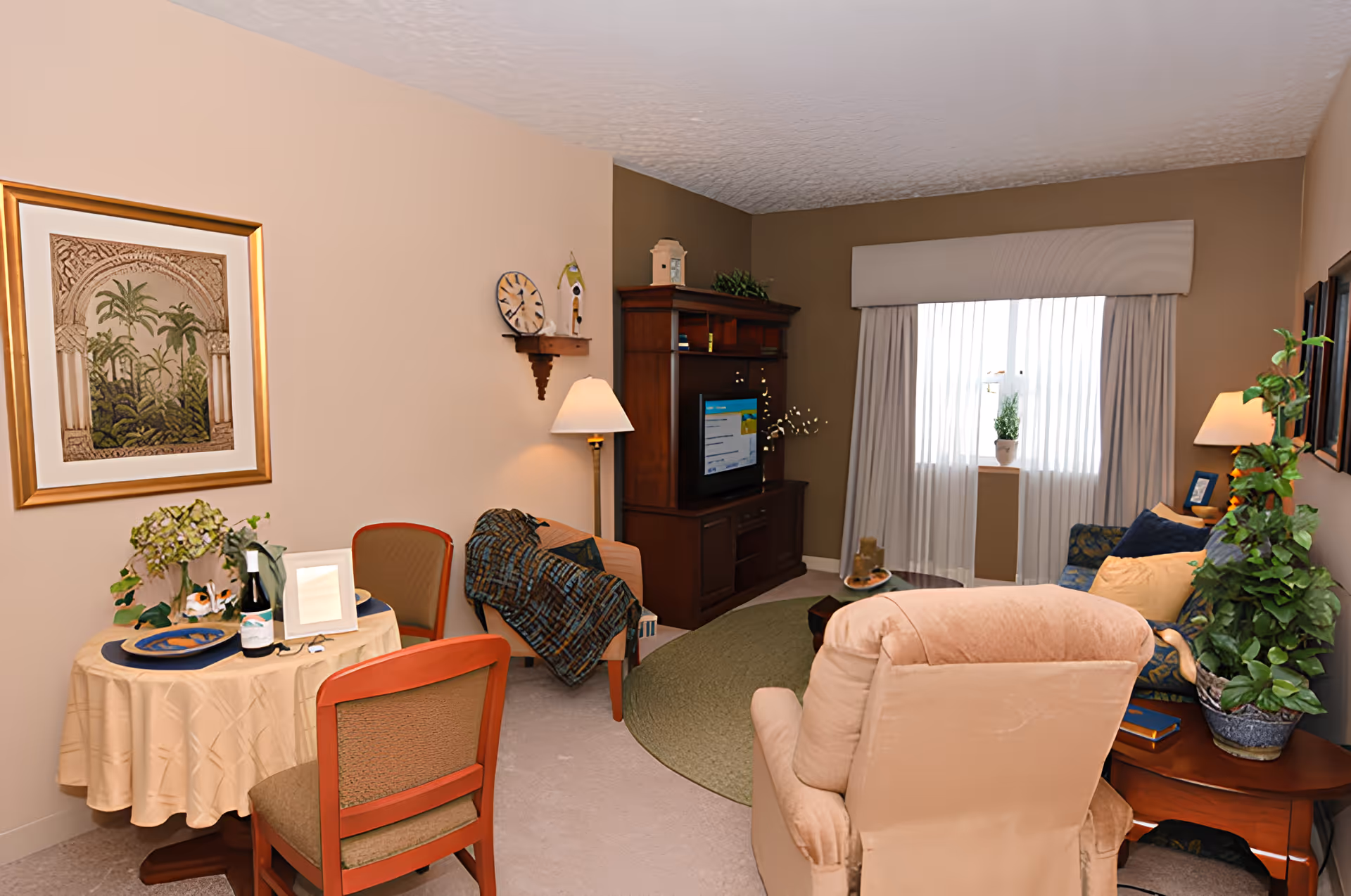 A cozy living room in a senior living facility with a beige recliner chair, a small round dining table set for two with a bottle of wine and flowers, a wooden entertainment center with a TV, a blue patterned sofa with cushions, a side table with a potted plant, and a window with white curtains letting in natural light.