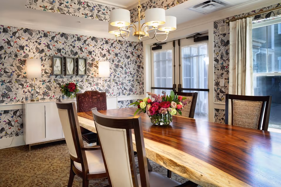 A dining room with a large wooden table featuring a natural edge and a floral centerpiece. The room has four chairs with cushioned seats and backs. The walls are decorated with butterfly-patterned wallpaper and two table lamps on a white sideboard. There are double glass doors and a window with light-colored curtains allowing natural light into the room. A modern chandelier with three lamps hangs above the table.