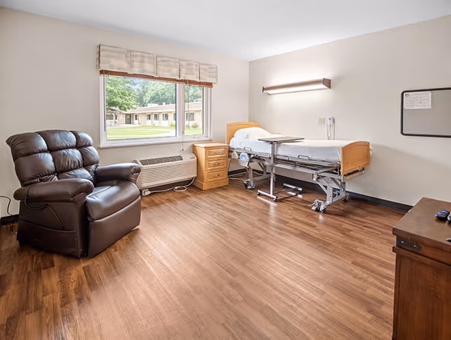 A senior living facility room with a hospital-style bed, a wooden nightstand, a large brown leather recliner chair, a window with a view of the outdoor grounds, and wood flooring. The room has neutral-colored walls and a wall-mounted light above the bed.