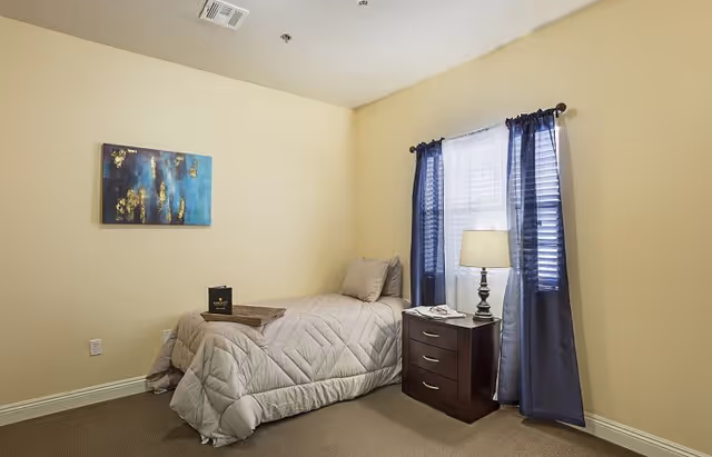 A small bedroom with beige walls and carpet, featuring a single bed with a gray quilt and pillows. Next to the bed is a dark wooden nightstand with three drawers, a lamp, and a telephone. A window with white blinds and dark blue curtains is behind the nightstand. A blue and gold abstract painting hangs on the wall above the bed.