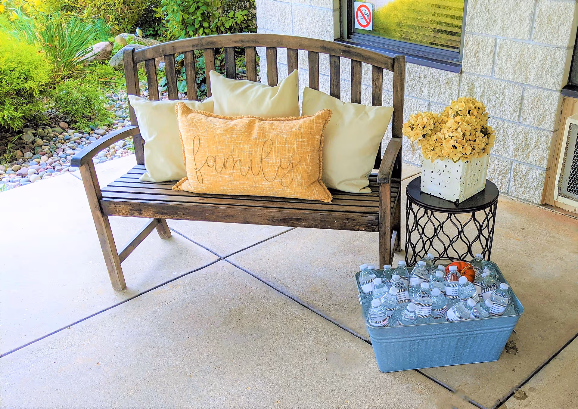 Wooden bench with decorative pillows including a yellow 'family' pillow on an outdoor patio next to a small table with flowers and a tub of bottled water.