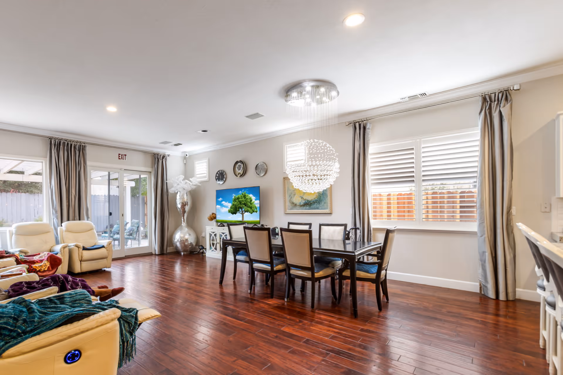 Open, well-lit senior living common room with reclining chairs, a dining table and chandelier, a TV, and large windows with curtains.