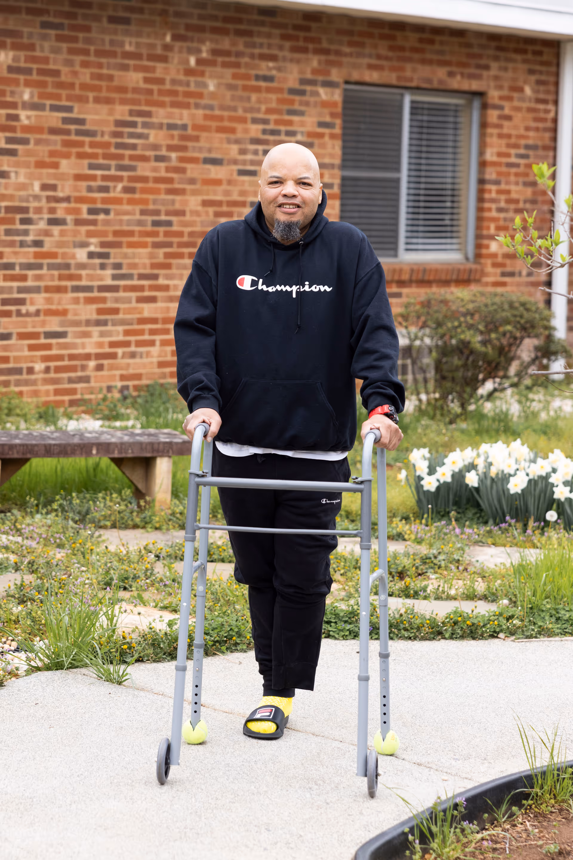 A man wearing a black Champion hoodie and black pants uses a walker while standing on a paved path outside a brick building with a window. There are flowers and greenery around the path.
