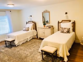 Bright bedroom with two twin beds, matching headboards, a dresser with mirror between them, two small benches, and a large area rug.