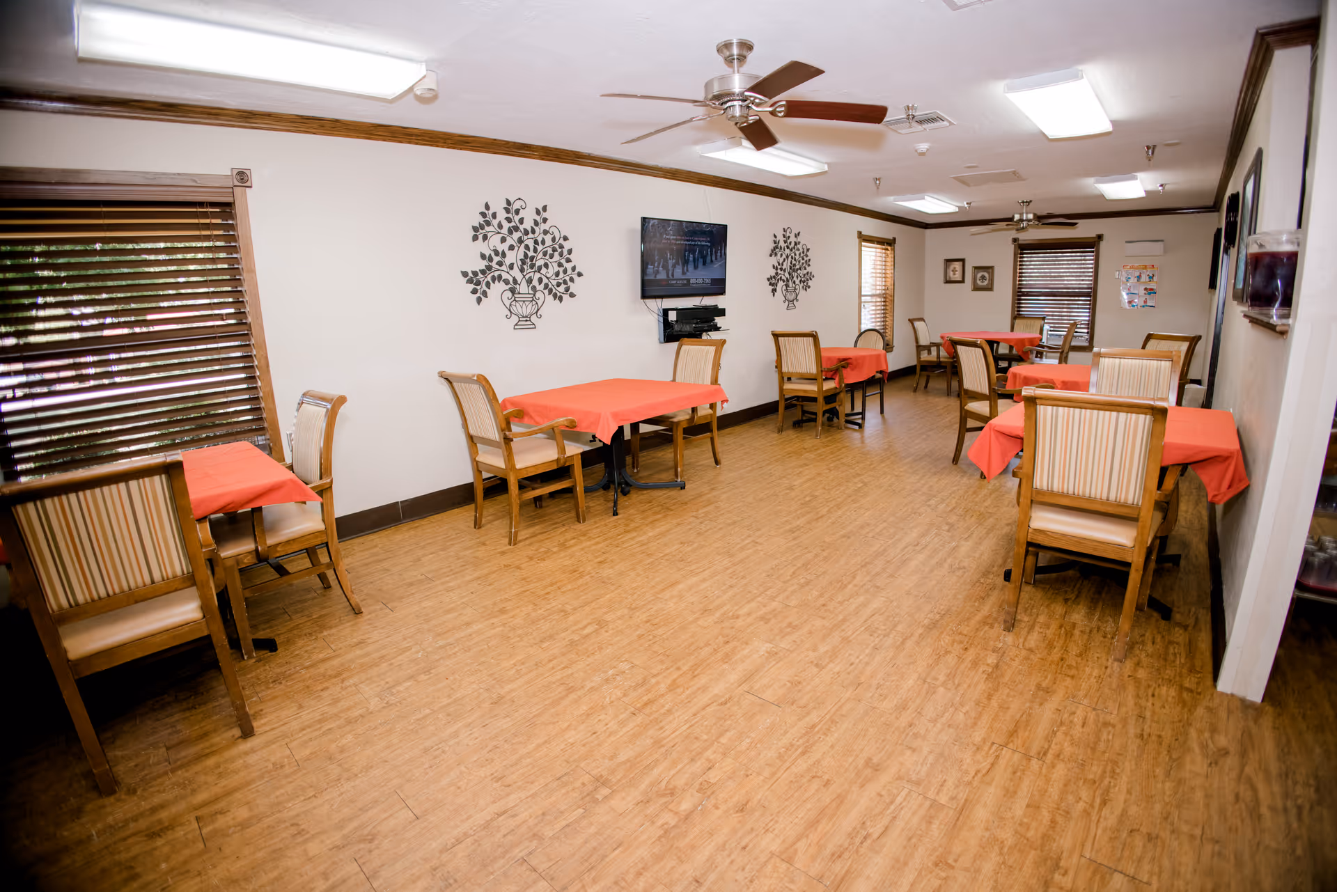 A dining room with several wooden tables covered with red tablecloths and wooden chairs with striped cushions. The room has wood flooring, white walls with decorative wall art, a ceiling fan, and a wall-mounted television. Windows with wooden blinds are visible on the left and at the far end of the room.