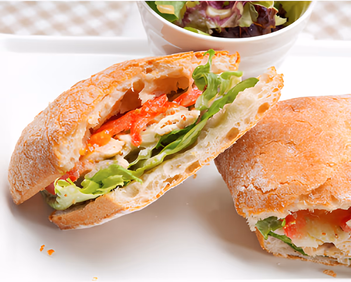 Close-up of a sandwich cut in half with lettuce, red peppers, and chicken on a white plate, accompanied by a bowl of mixed green salad in the background.