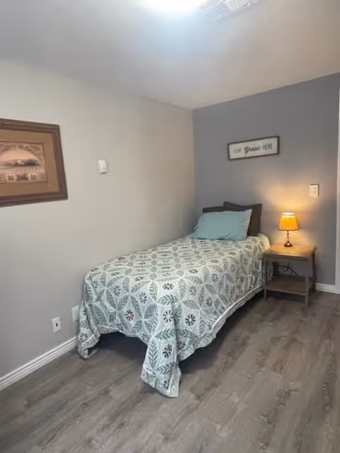 Small bedroom with a single bed covered in a patterned quilt, a nightstand with a lit lamp, wall art, and wood-look flooring.