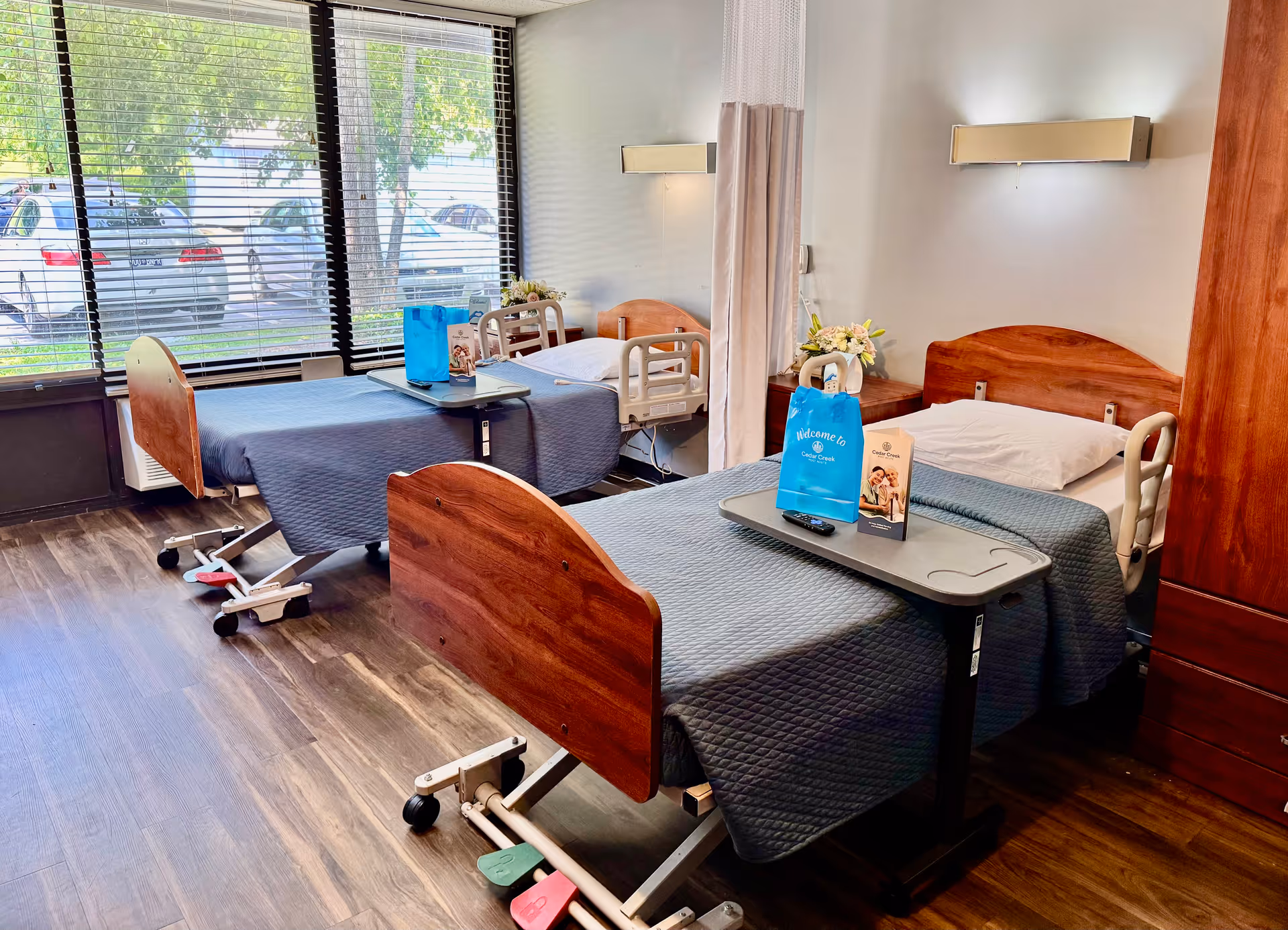 Shared patient room with two adjustable hospital beds covered in blue quilts, overbed tables, and a large window with blinds.