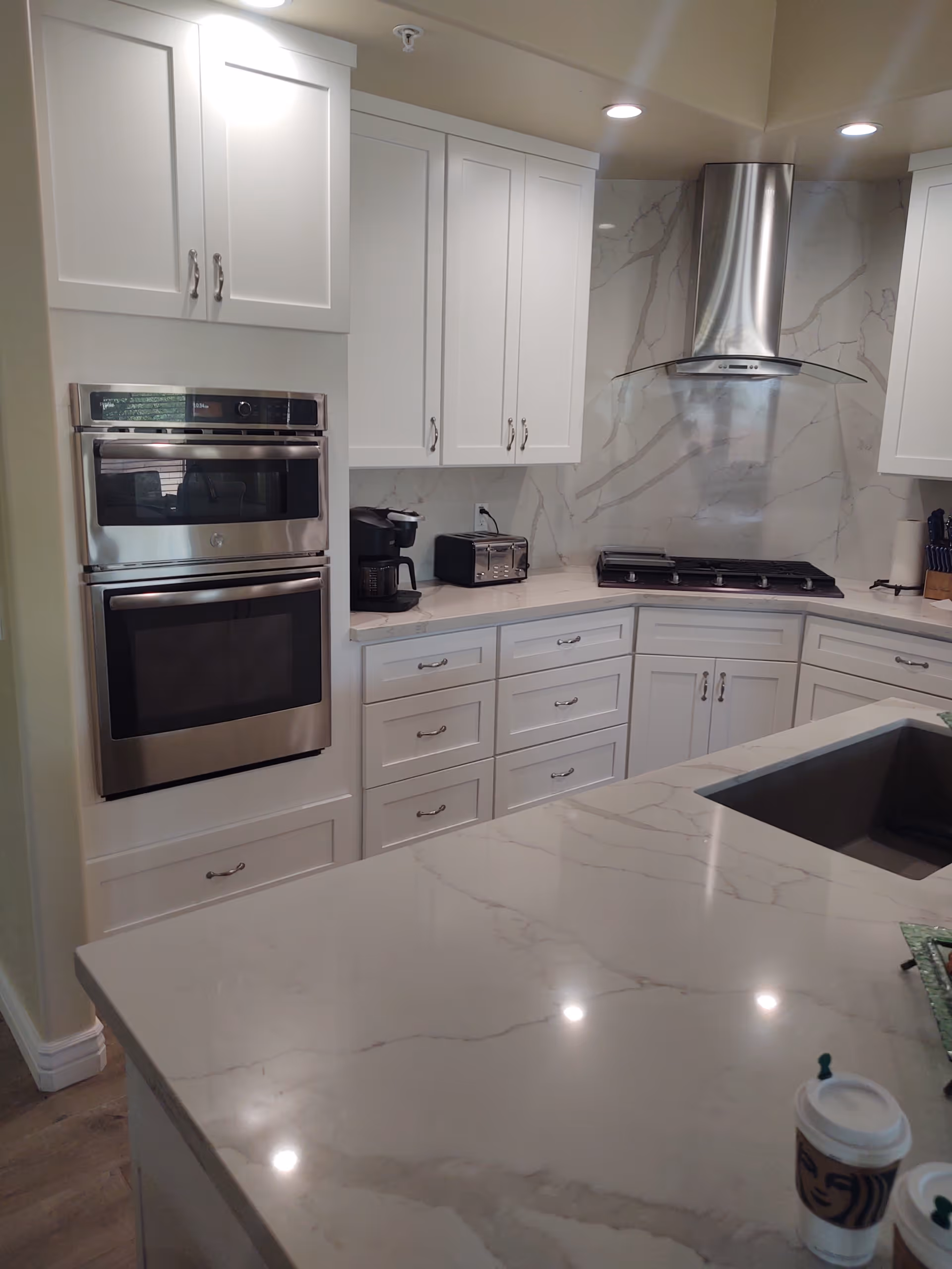 Modern kitchen with white cabinets, marble countertops, a built-in stainless steel double oven, a coffee maker, a toaster, a gas stove with a stainless steel range hood, and a sink on the island countertop. Two Starbucks coffee cups are visible on the island.