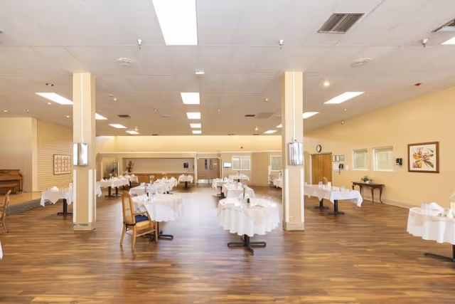 Spacious dining room with round tables set with white tablecloths, wooden chairs, and wood-look flooring under overhead lights.