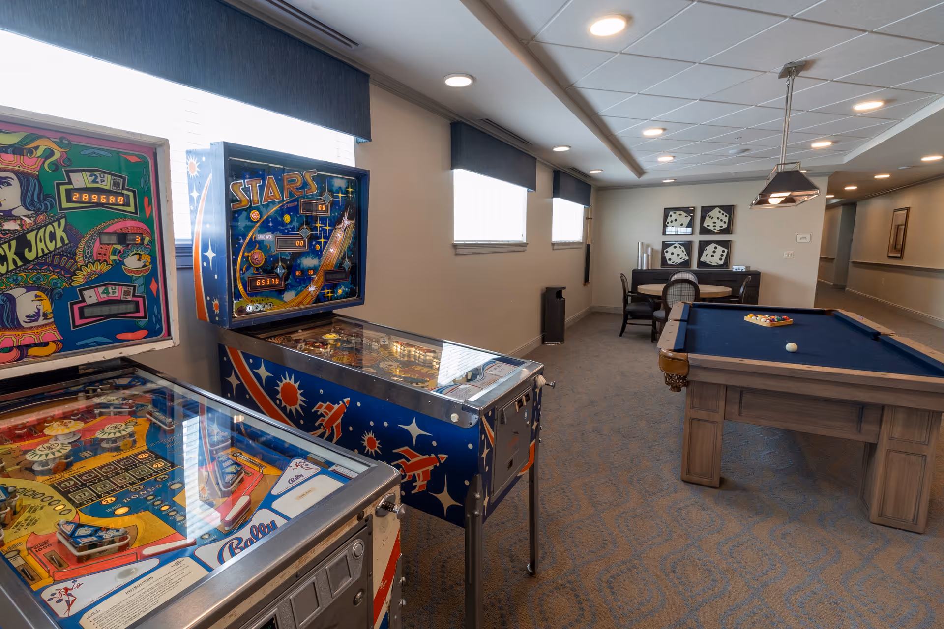 Recreation room interior with pinball machines, a pool table, and a small seating area under ceiling lights.